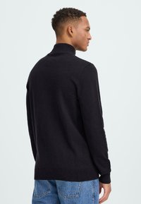 Juodas megztinis su aukštu kaklu, pagamintas iš minkštos medžiagos. Prigludusi konstrukcija su ilgomis rankovėmis ir pylimo rankovėmis, derinama su šviesiai mėlynomis džinsomis.