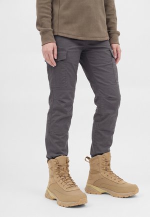 Personne portant un pantalon cargo gris et des bottes de randonnée montantes couleur beige, debout sur un fond clair uni.