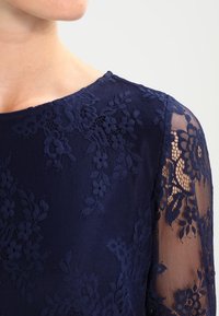 Abito in pizzo blu navy con motivi floreali e maniche lunghe trasparenti. Il collo è rotondo, con una texture liscia che mette in risalto dettagli intricati.