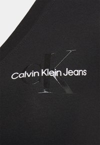 Svart tygtank topp med en glänsande Calvin Klein Jeans-logotyp i vitt och svart. Slät textur med en åtsittande passform och rena sömmar.