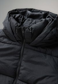 Giacca imbottita nera con collo alto, chiusura con zip e tessuto leggero e strutturato con finitura leggermente stropicciata.