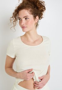 Top blanc en tricot côtelé à manches courtes avec un col rond, présentant une texture subtile et un ourlet légèrement court. Le modèle tient le tissu.