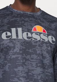 Szara koszulka z nadrukiem w kamuflaż, z okrągłym dekoltem. Na przodzie znajduje się logo "ellesse" w białym kolorze oraz wielobarwne logo w kształcie łuku w kolorze pomarańczowym i czerwonym.