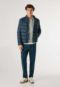 Mornarsko modra puffer jakna s horizontalnim šivom, v paru s svetlo sivo kapuco, temno modrimi hlačami in belimi supergami s bež podplati.