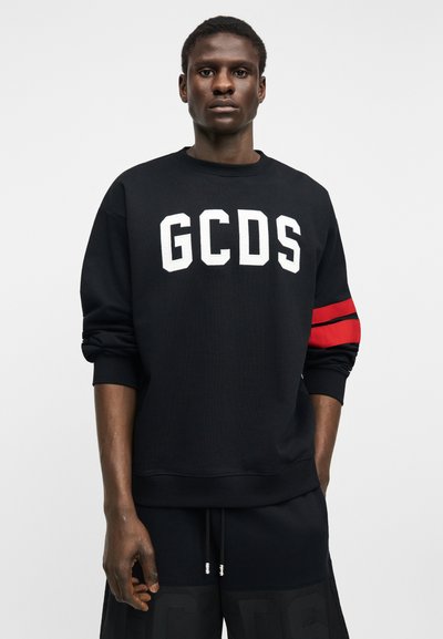 Άνδρας που φοράει μαύρο φούτερ με λευκά γράμματα «GCDS» και κόκκινες ρίγες στο αριστερό μανίκι, συνδυασμένο με μαύρο σορτς, στέκεται μπροστά σε απλό φόντο.