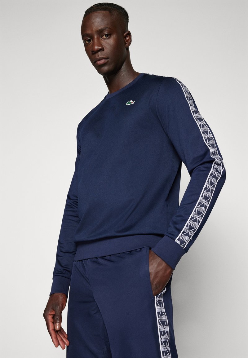 Sweatshirt blu navy con maniche lunghe, caratterizzato da un motivo geometrico bianco testurizzato lungo i lati e un piccolo logo verde sul petto.