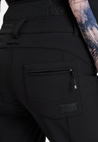 Zwarte sportbroek met een ritszak, textuurstof en een logo patch. Het ontwerp heeft gestikte details en een getailleerd silhouet.