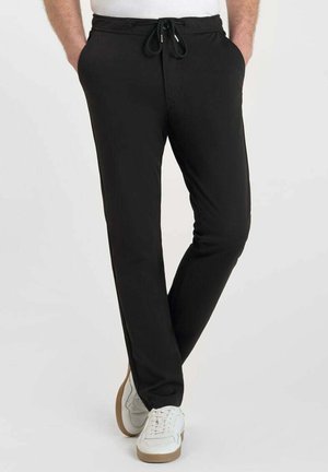 Man in zwarte jogger-pantalons met zijn handen in de zakken en witte sneakers met bruine zolen, staand met gekruiste benen.