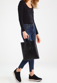 Svart läder tote-väska med präglat "DKNY"-logotyp, rektangulär form, två korta handtag, slät textur, matchar med denim och svarta sneakers.