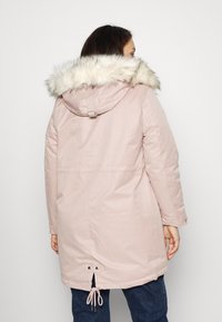 Eine Frau trägt eine hellrosa Parka mit einer mit Pelz gefütterten Kapuze. Sie wird von hinten gesehen, während sie vor einem schlichten weißen Hintergrund steht.