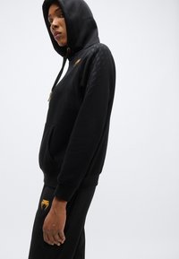 Hoodie noir avec un motif texturé et une poche kangourou, arborant un logo doré sur la poitrine. Associé à un pantalon de jogging noir assorti.