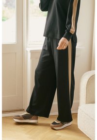 Osoba ubrana w czarne szerokie spodnie i dopasowany top z beżowymi paskami po bokach, spacerująca wewnątrz pomieszczenia w pobliżu okna i kremowego fotela.