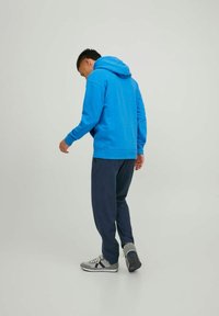 Blauwe hoodie met een gestructureerde afwerking, gecombineerd met donkere broeken en grijze sneakers. De hoodie heeft een trekkoord en een kangoeroezak.