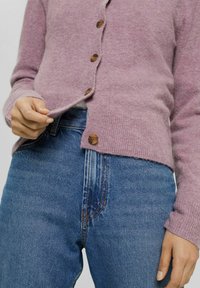 Person med en rosa knappad kofta och blå jeans, som håller i kanten av koftan vid midjan.