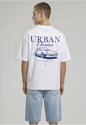 Jeune homme portant un t-shirt blanc avec une voiture classique bleue et le texte "Urban Classics", associé à un short en jean bleu clair.