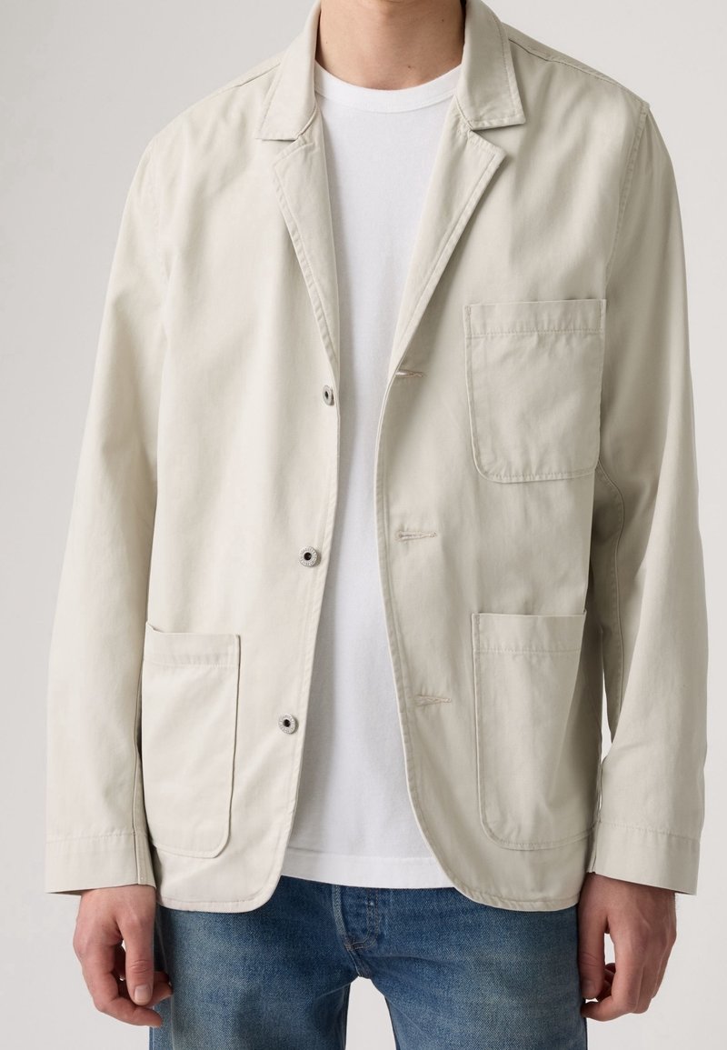 Homme portant un blazer décontracté beige clair sur un T-shirt blanc et un jean bleu, debout devant un fond uni.