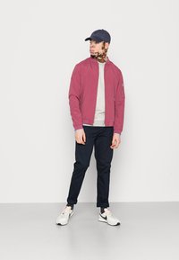 Lett, maroon bomberjakke med stående krage, grå genser, marineblå bukser og hvite sneakers. Kappe med kort brem i marineblå.