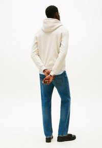 Felpa beige con cappuccio e una texture morbida, abbinata a jeans blu a gamba dritta e scarpe nere, vista da dietro con le mani incrociate.