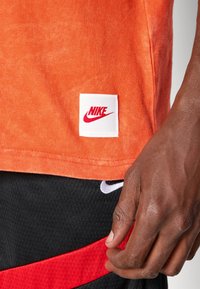 Orange T-shirt lavet af blød stof med et hvidt Nike-logo patch med rød tekst, parret med sorte shorts med røde og hvide accenter.