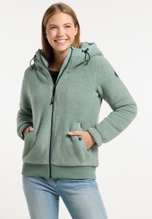 Lachende vrouw die een lichtgroene fleece hoodie met rits en capuchon draagt, met haar handen in de zakken, gecombineerd met lichtblauwe spijkerbroek.