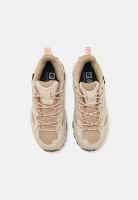 Beige utendørssko laget av nett og semsket lær, med teksturerte detaljer, oransje og hvite snørebånd, og en svart logo på tungen.