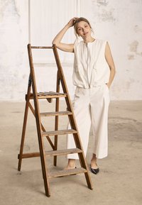 Femme en gilet blanc sans manches et pantalon, debout à côté d'une échelle en bois dans une pièce lumineuse et vide avec des murs texturés.