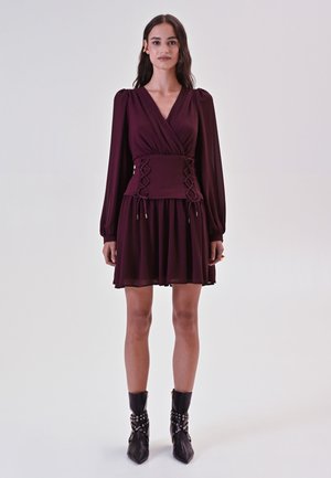 Burgunderfarbendes Wickelkleid mit langen Puffärmeln, geraffter Taille und dekorativen Schnürdetails, kombiniert mit schwarzen Knöchelstiefeln mit silbernen Akzenten.