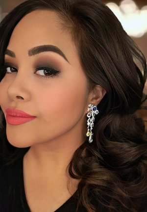 Frau mit dunklen, gelockten Haaren, die funkelnde, hängende Ohrringe und dunkles Augen-Make-up trägt, posiert mit leichtem Lächeln vor unscharfem Hintergrund.