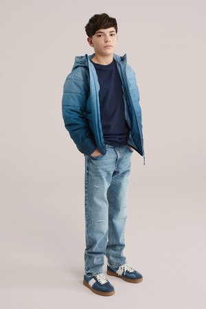 Dreng står med hænderne i lommerne iført en blå dynejakke, mørkeblå skjorte, lyseblå jeans og blå sneakers med hvide striber.