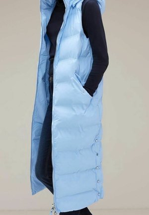 Person trägt ärmellose hellblaue Steppweste über schwarzem Langarmshirt und dunklen Jeans, mit Hand in der Westentasche.