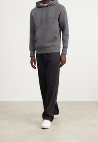 Šedá mikina s kapucí, přední kapsou a stahovací šňůrkou, v kombinaci s černými širokými kalhotami a bílými teniskami. Minimalistické logo na mikině.