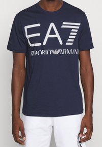 Navy blue cotton t-shirt with short sleeves. Large white print displaying "EA7" and "Emporio Armani" across the chest. Simple design, crew neck.