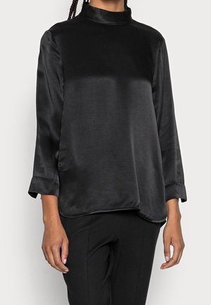 Kvinde iført en sort satinbluse med lange ærmer, høj hals og manchetter, kombineret med sorte bukser mod en ensfarvet baggrund.