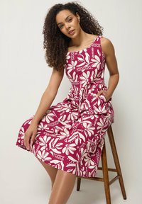 Mujer con cabello rizado sentada en un taburete de madera, vistiendo un vestido sin mangas, de longitud hasta la rodilla, con estampado floral rojo y blanco y bolsillos.