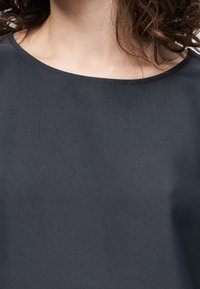 Femme aux cheveux bouclés foncés portant un haut gris foncé uni avec un large décolleté arrondi, visible des épaules à la partie supérieure de la poitrine.