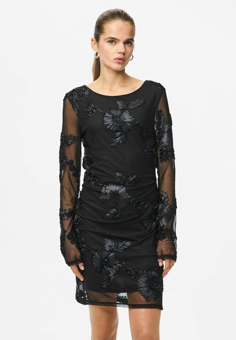 Czarna sukienka z długimi, przezroczystymi rękawami, ozdobiona haftowanymi kwiatowymi wzorami. Posiada dopasowaną sylwetkę i okrągły dekolt.