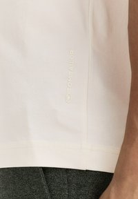 Tricou din bumbac bej deschis, cu un eticheta logo subtilă pe cusătura laterală, având o textură netedă și un design cu o croială lejeră.