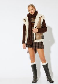 Gilet di pelliccia sintetica marrone con una fodera beige, maglione a fantasia, gonna a quadri e stivali alti neri con calze bianche.