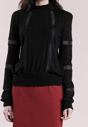 Nainen, joka käyttää mustaa teksturoitua pitkähihaista yläosaa läpikuultavilla paneeleilla ja korkealla röyhelöidyllä kauluksella, yhdistettynä punaiseen hameeseen.