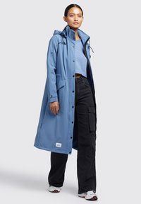 Abrigo largo azul impermeable con capucha, botones a presión en el frente y bolsillos laterales, combinado con pantalones cargo negros y zapatos deportivos.