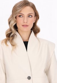 Beige kunstlæderjakke med en struktureret krave, der har en enkelt knaplukning, glat tekstur og minimalistisk design.