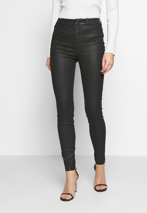 Schwarze Faux-Leder Skinny-Hosen mit hoher Taille, strukturiertem Finish und Reißverschlüssen an den Knöcheln, ergänzt durch schwarze Stiletto-Sandalen.