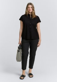 Chemise noire à manches courtes, à bouton, coupe décontractée, associée à un pantalon noir ajusté, des sandales tressées noires et un sac fourre-tout à rayures noir et blanc.