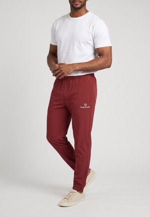 Hombre con camiseta blanca, pantalones deportivos rojos Sergio Tacchini y zapatillas beige, de pie contra un fondo gris liso.