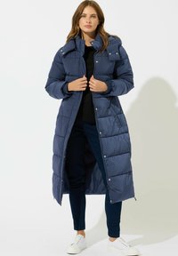 Abrigo acolchado largo de color azul marino con capucha, con cierre delantero de cremallera, botones a presión y diseño acolchado. Combinado con pantalones oscuros y zapatillas blancas.