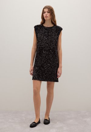 Frau steht vor einer schlichten Wand und trägt ein ärmelloses schwarzes Pailletten-Kleid und schwarze, bestickte, flache Schuhe.