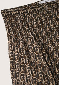 Tissu noir avec un motif répété de logo "MNG" beige, montrant un gros plan de la ceinture plissée d'une jupe et de son étiquette.