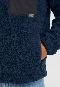 Marineblauer Fleece-Pullover mit einer strukturierten, plüschigen Außenseite, einer schwarzen Nylon-Brusttasche und seitlichem Reißverschluss. Mit einem Logo-Label auf der Tasche.