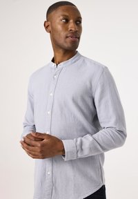 Camisa de botones azul claro con cuello mao, que presenta una tela texturizada y mangas largas. Los botones son blancos y el corte es casual.
