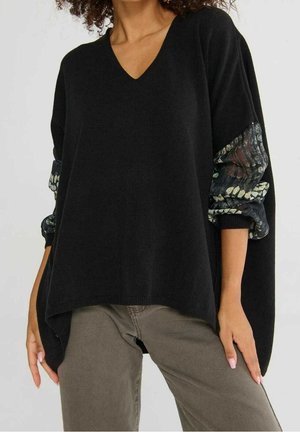 Femme portant un pull ample noir à col en V avec des manches transparentes à motifs, associée à un pantalon vert olive, debout avec les mains détendues.